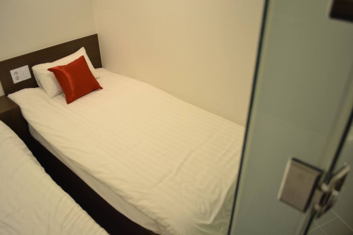 A single bed with white bedding and a red pillow, a wooden headboard, and a light-colored wall.