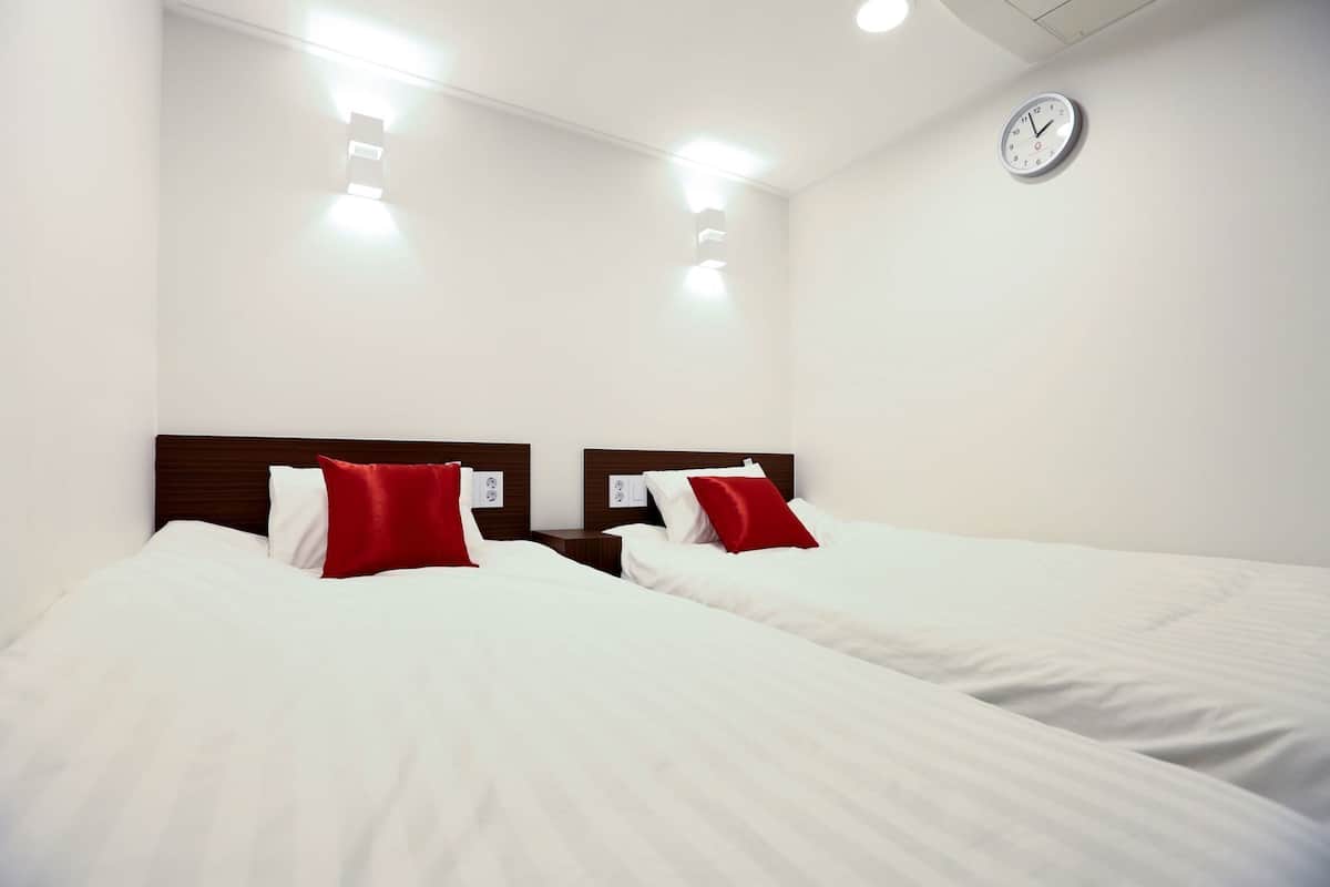 A hotel room with two single beds, white bedding, red pillows, a wooden headboard, a wall clock, and recessed ceiling lighting.