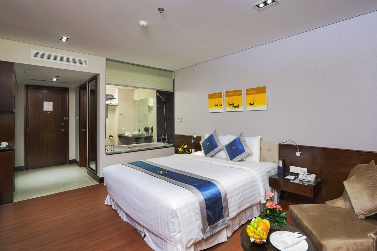 Une chambre d’hôtel avec un grand lit, une table de chevet avec un téléphone et une lampe, une chaise, et une petite table avec des fruits et des fleurs.