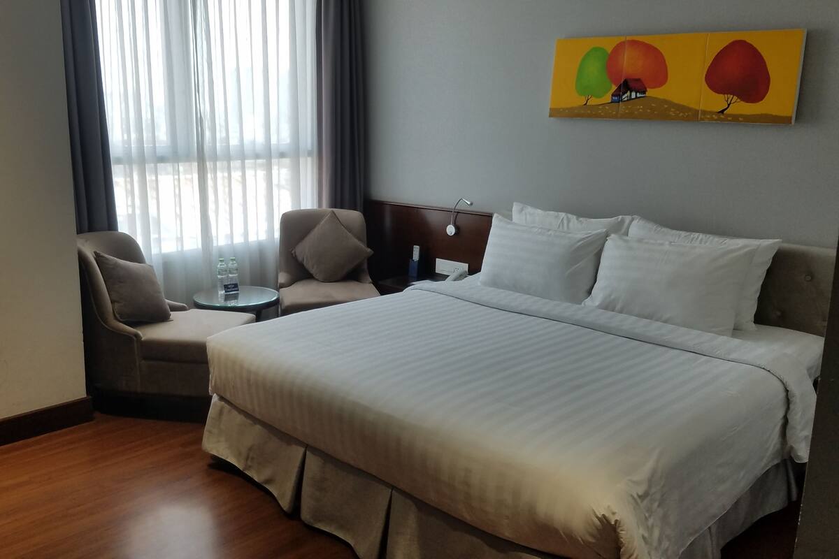 Hotelkamer met een groot bed, een stoel, een klein tafeltje met waterflesjes en een schilderij aan de muur.