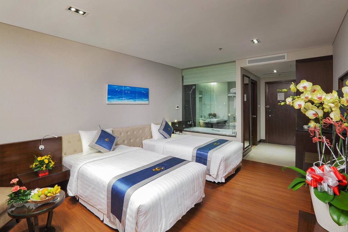 Een hotelkamer met twee bedden, een klein tafeltje met fruit en bloemen, een schilderij aan de muur en een badkamer zichtbaar door een glazen deur.