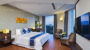 Een moderne hotelkamer met een groot bed, een zithoek met een fauteuil en een tafeltje, een groot raam met uitzicht op de stad, en een schilderij aan de muur.