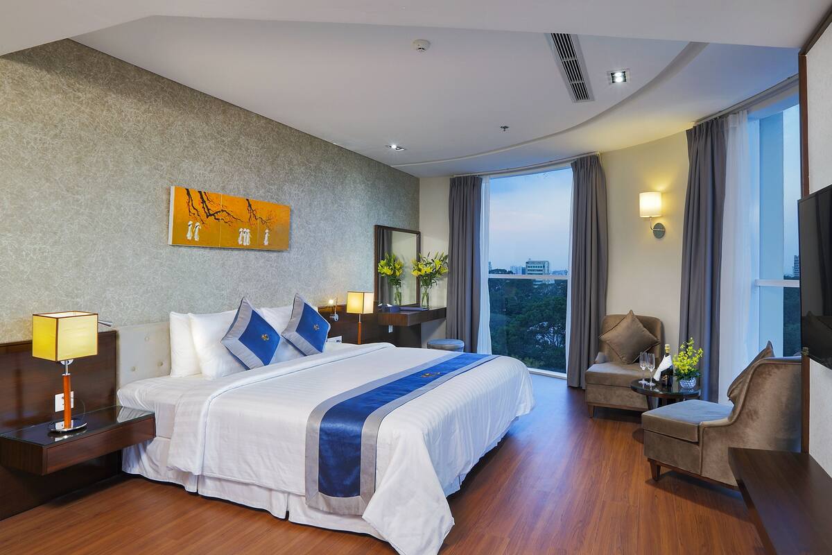 Een moderne hotelkamer met een groot bed, een zithoek met een fauteuil en een tafeltje, een groot raam met uitzicht op de stad, en een schilderij aan de muur.