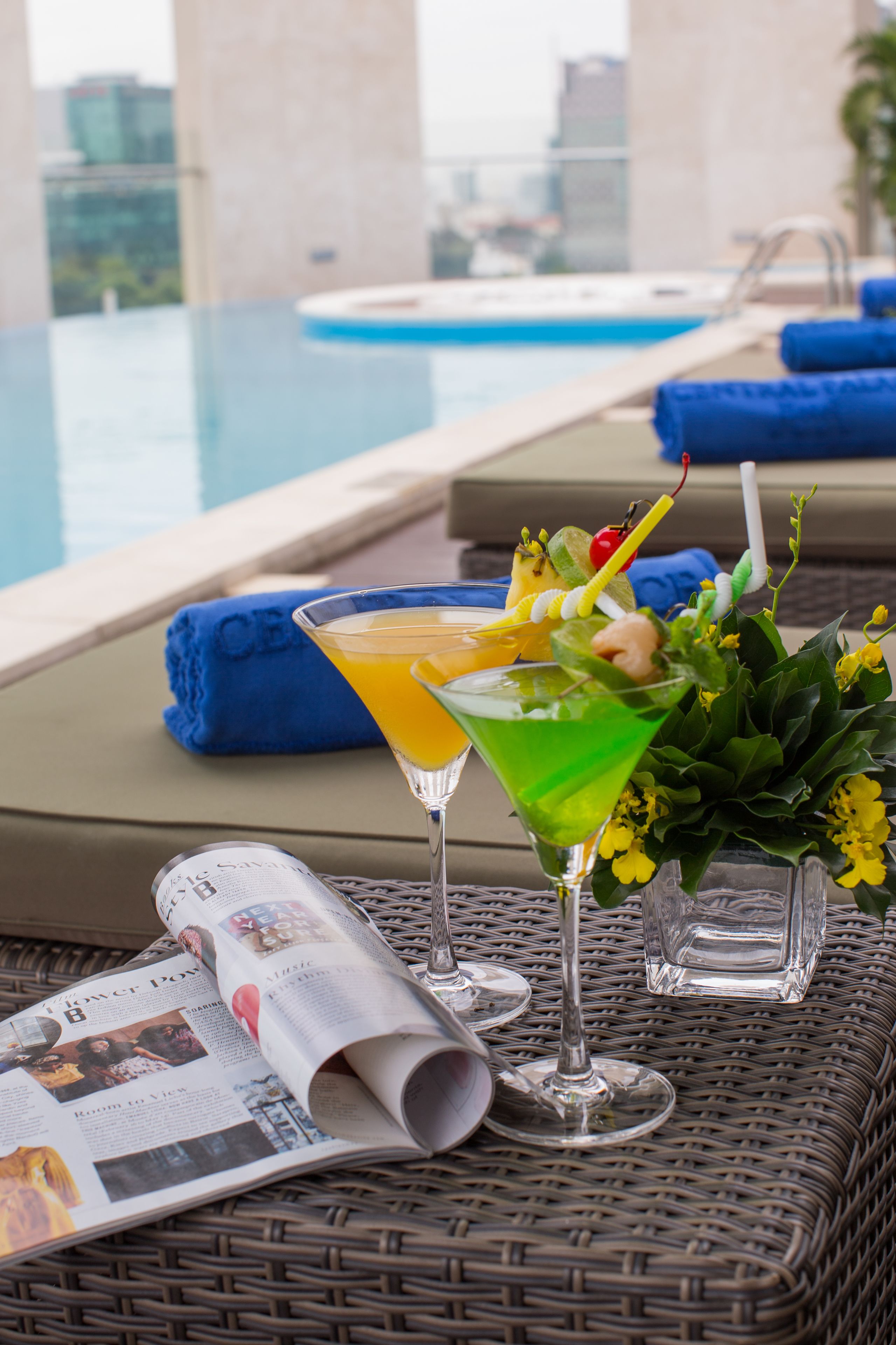 A poolside setting with two cocktails, a magazine, and a view of the pool.