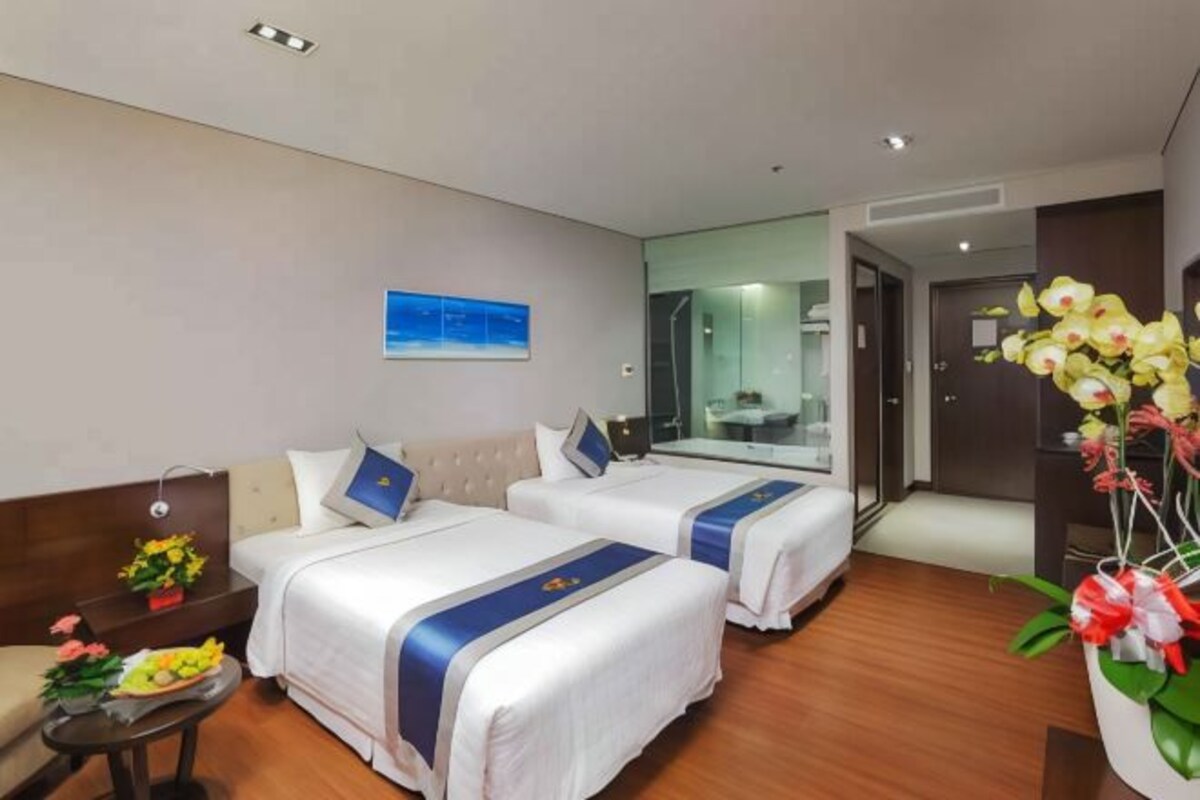 A hotel room with two beds, a sofa, a small table with flowers, a mirror, and a view of the bathroom.