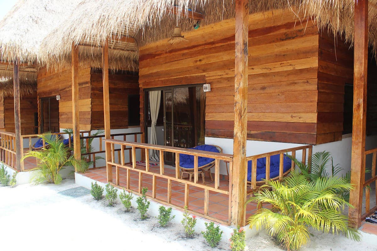 A wooden cabin with a thatched roof, a covered porch with seating, and small plants.