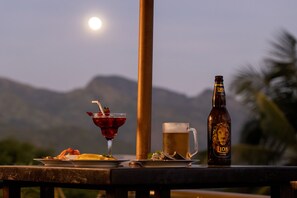 A table with a beer bottle, a glass of red drink, and a mug, set against a backdrop of mountains and a moon. - The Albatross, Kandy (Maberiyatenna)