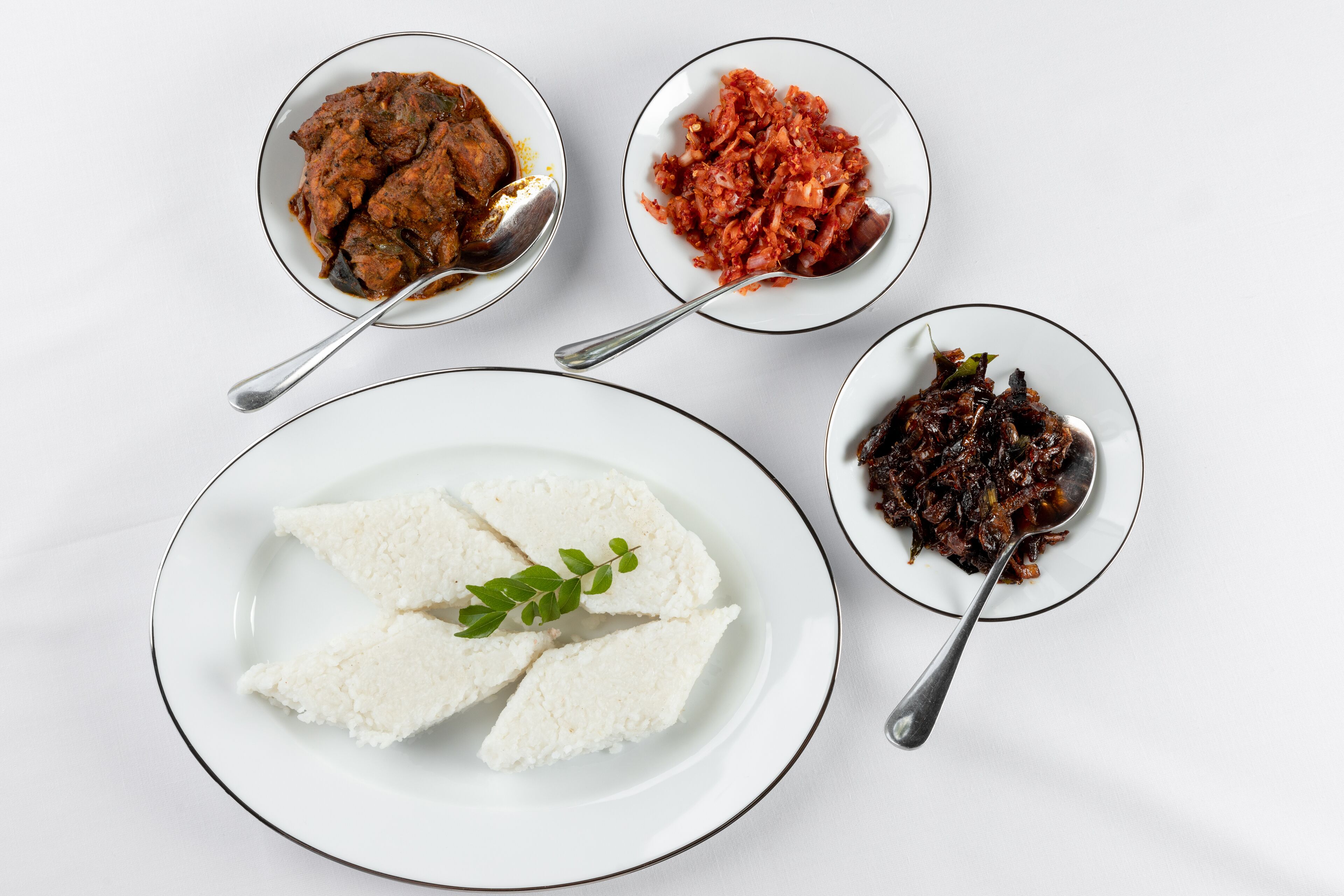 Una mesa con cuatro platos: un bol de arroz blanco, un bol de estofado oscuro, un bol de chutney rojo y un bol de comida frita oscura.