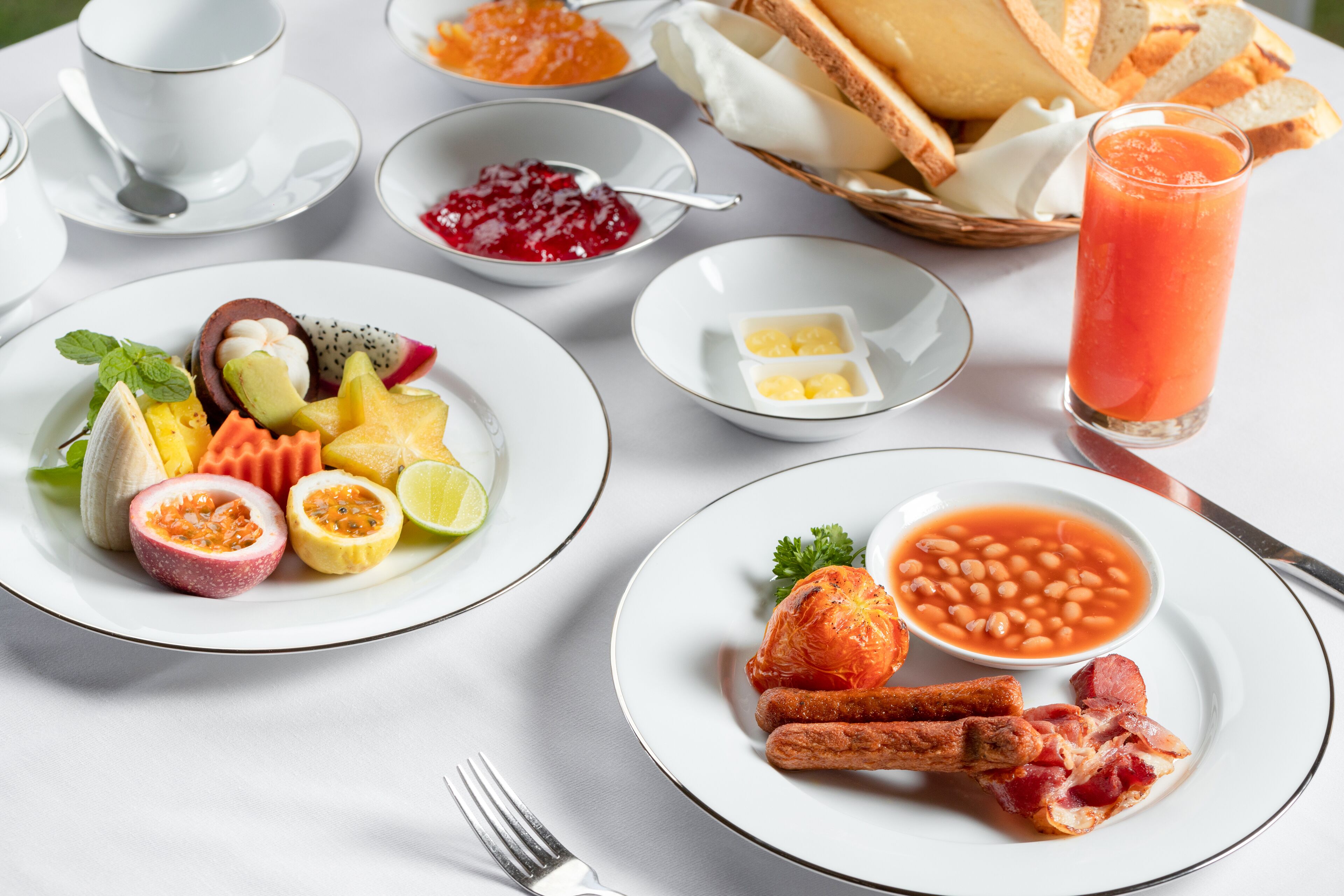 A breakfast setting with plates of fruit, toast, beans, and bacon.