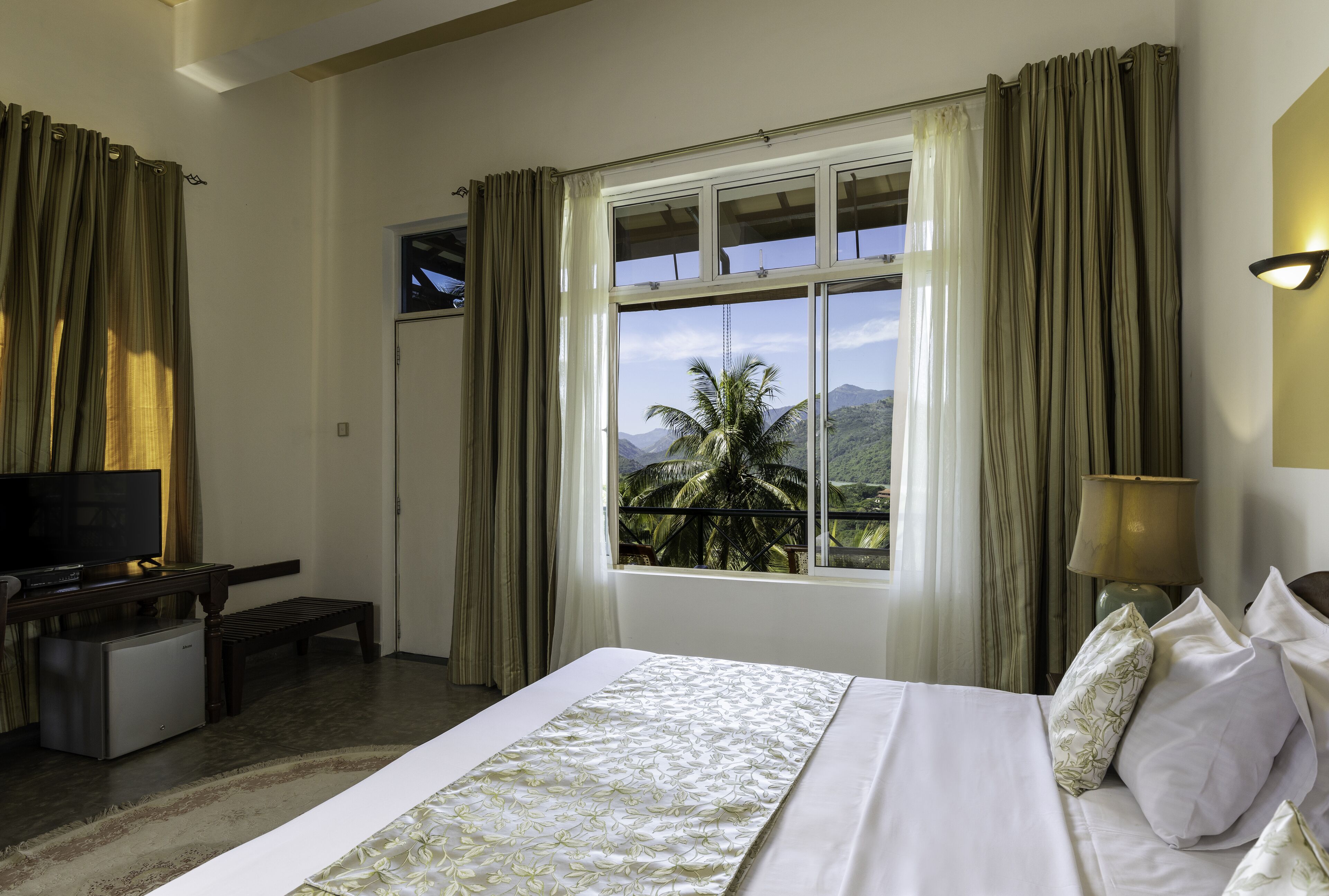 Una habitación de hotel con cama, televisor, escritorio con computadora y ventana con cortinas.