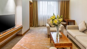 Una habitación de hotel moderna con una televisión de pantalla plana, una mesa de centro de madera, un sofá color crema y un ventanal con cortinas.