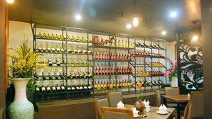 A restaurant interior with multiple dining tables and chairs, a wine rack with various bottles, and a large vase with flowers.