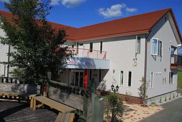 A two-story building with a red roof and a sign that reads "Blane Flowers." There is a wooden deck with benches in front. - Petit Hotel Blanc Fleur (Nakafurano)