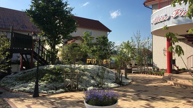 A hotel courtyard with a paved walkway, a large planter with flowers, and a building with a red roof and a sign reading "The Blonde Hotel". - Petit Hotel Blanc Fleur (Nakafurano)
