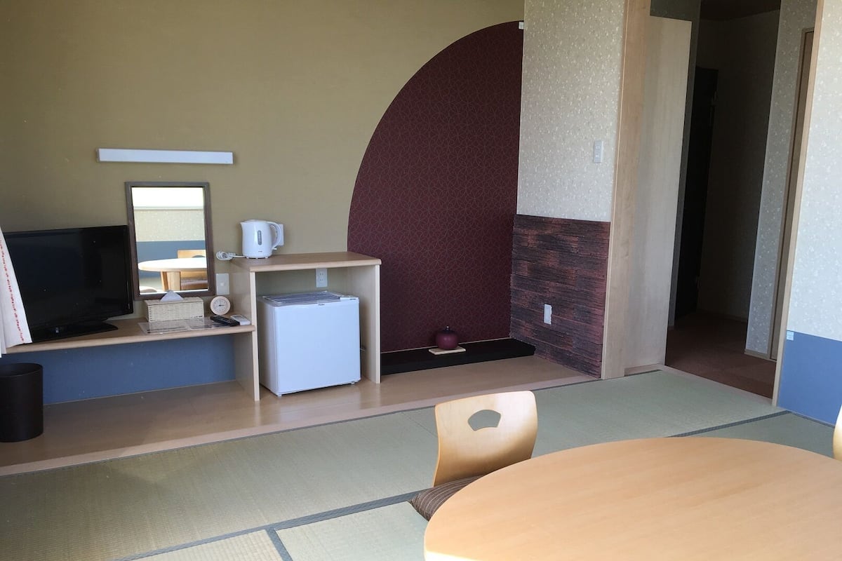 Een kamer met een televisie, een kleine koelkast, een waterkoker, een klok en een ronde tafel met stoelen.
