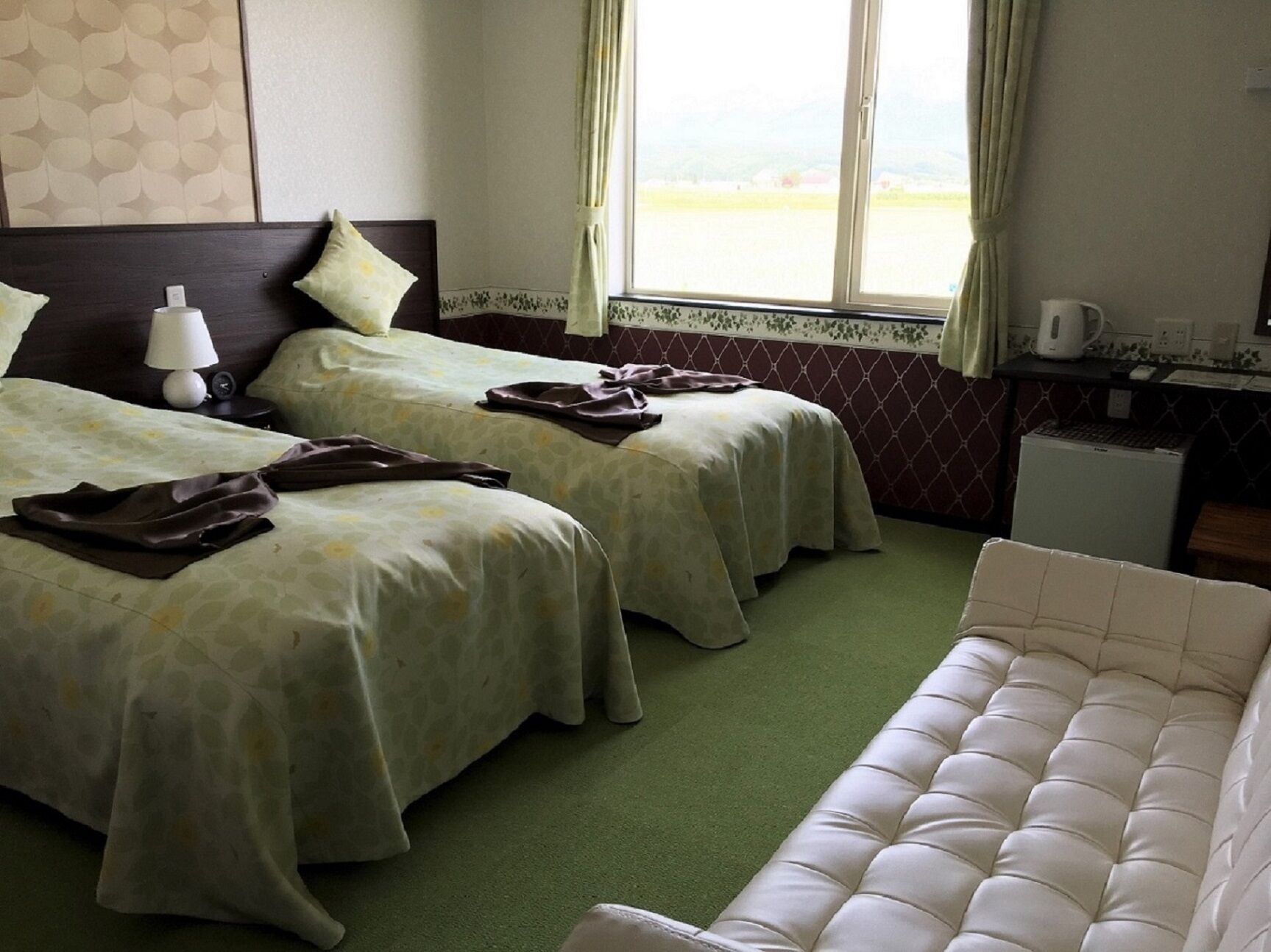 Une chambre d’hôtel avec deux lits, une moquette verte, un canapé blanc, un petit bureau avec un ordinateur et une fenêtre avec des rideaux.