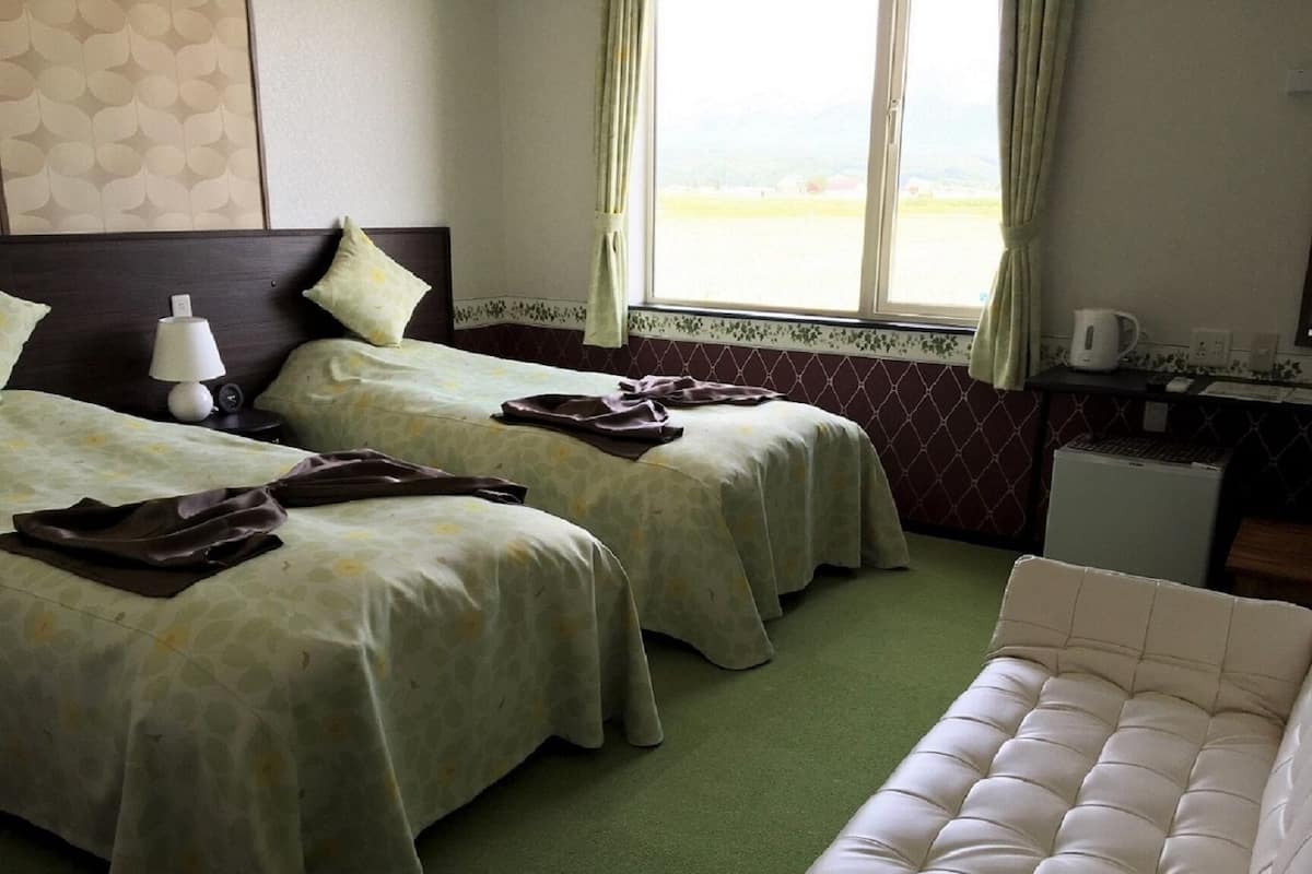Een hotelkamer met twee bedden, een groen tapijt, een witte bank, een klein bureau met een computer, en een raam met gordijnen.