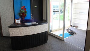 A reception desk with a framed picture, a vase with red flowers, and a view of a door leading outside. - Petit Hotel Blanc Fleur (Nakafurano)