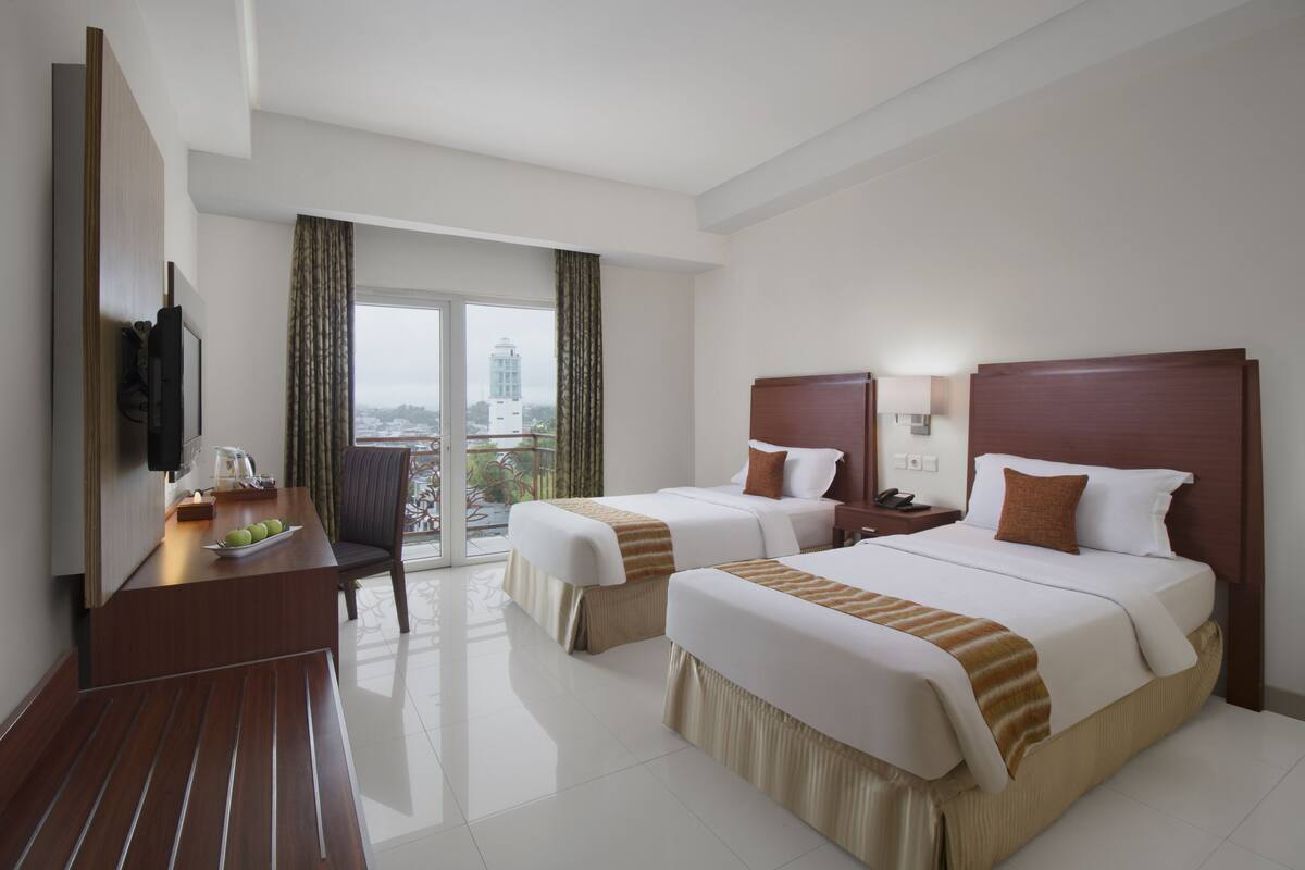 Ein Hotelzimmer mit zwei Betten, einem Schreibtisch mit Fernseher und Blick auf eine Stadtlandschaft.