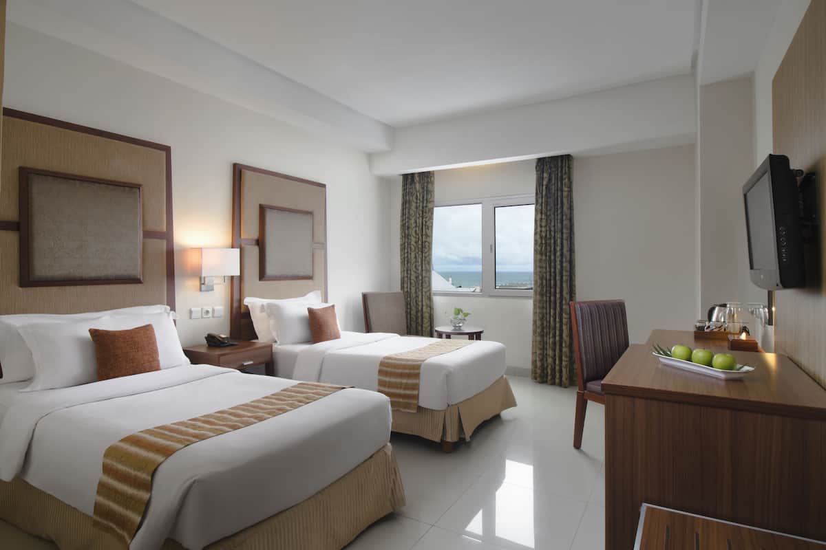 Ein Hotelzimmer mit zwei Betten, einem Schreibtisch mit Obstschale, einem Fernseher und Blick aufs Meer.