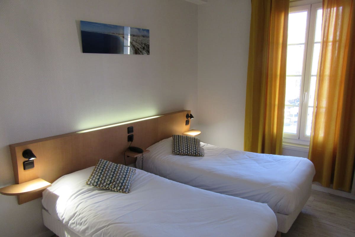 A hotel room with two beds, bedside lamps, a nightstand, a wall-mounted phone, and a window with curtains.