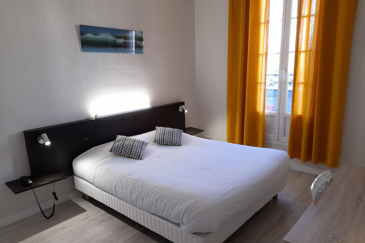 A neatly made bed with two patterned pillows, a bedside table with a lamp, and a window with yellow curtains.