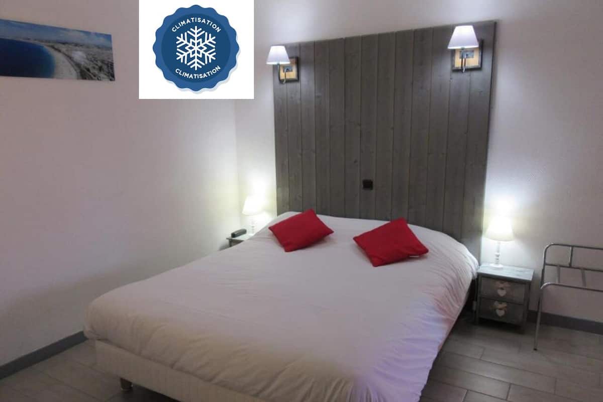 A hotel room with a large bed, two red pillows, a wooden headboard, bedside lamps, and a small bedside table.