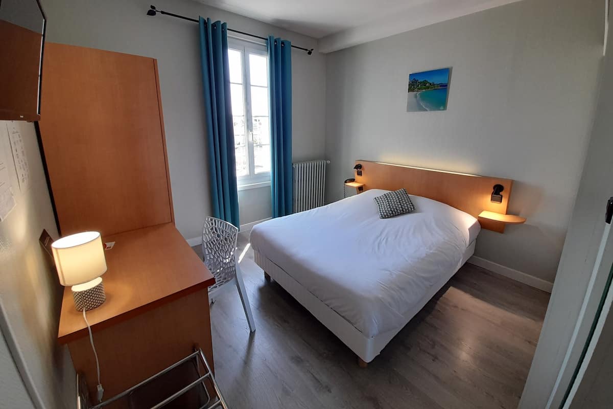 A hotel room with a bed, bedside lamps, a desk with a lamp, a chair, a window with curtains, and a painting on the wall.