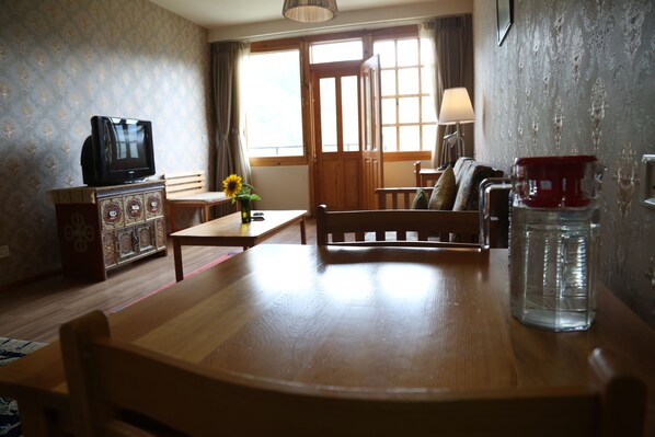 Una habitación con una mesa de madera, un televisor en un mueble bajo, un sofá, una lámpara y un jarrón con flores.