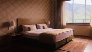 A hotel room with a large bed, a nightstand, a lamp, and a view of the outdoors through a window. - Bhutan Suites (Thimphu)