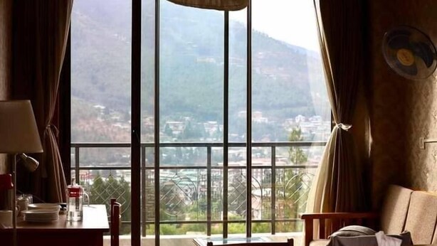 A room with a large window offering a view of a mountainous landscape. There is a table with a lamp and a chair. - Bhutan Suites (Thimphu)