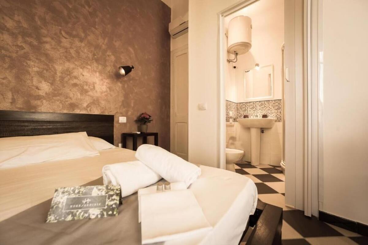 A hotel room with a checkered floor, a bed with neatly folded towels, a bedside table with a lamp, and a bathroom visible through an open door.