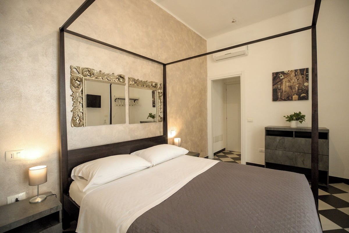 A bedroom with a four-poster bed, a large mirror, bedside lamps, a dresser, and a painting on the wall.