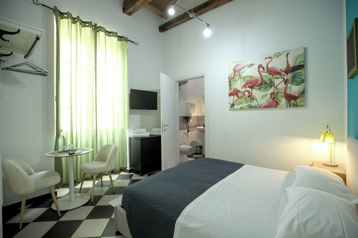 A hotel room with a checkered floor, a bed with white and grey bedding, a desk with a chair, a painting of flamingos, and a bathroom visible through an open door.