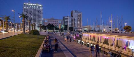 A waterfront promenade with a row of restaurants, palm trees, and a high-rise building in the background.