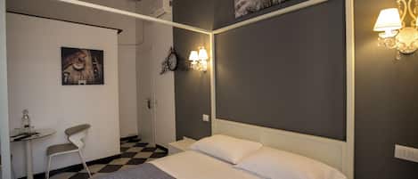 Un dormitorio con cama, una silla, una mesa con una botella y vasos, y lámparas fijadas a la pared.