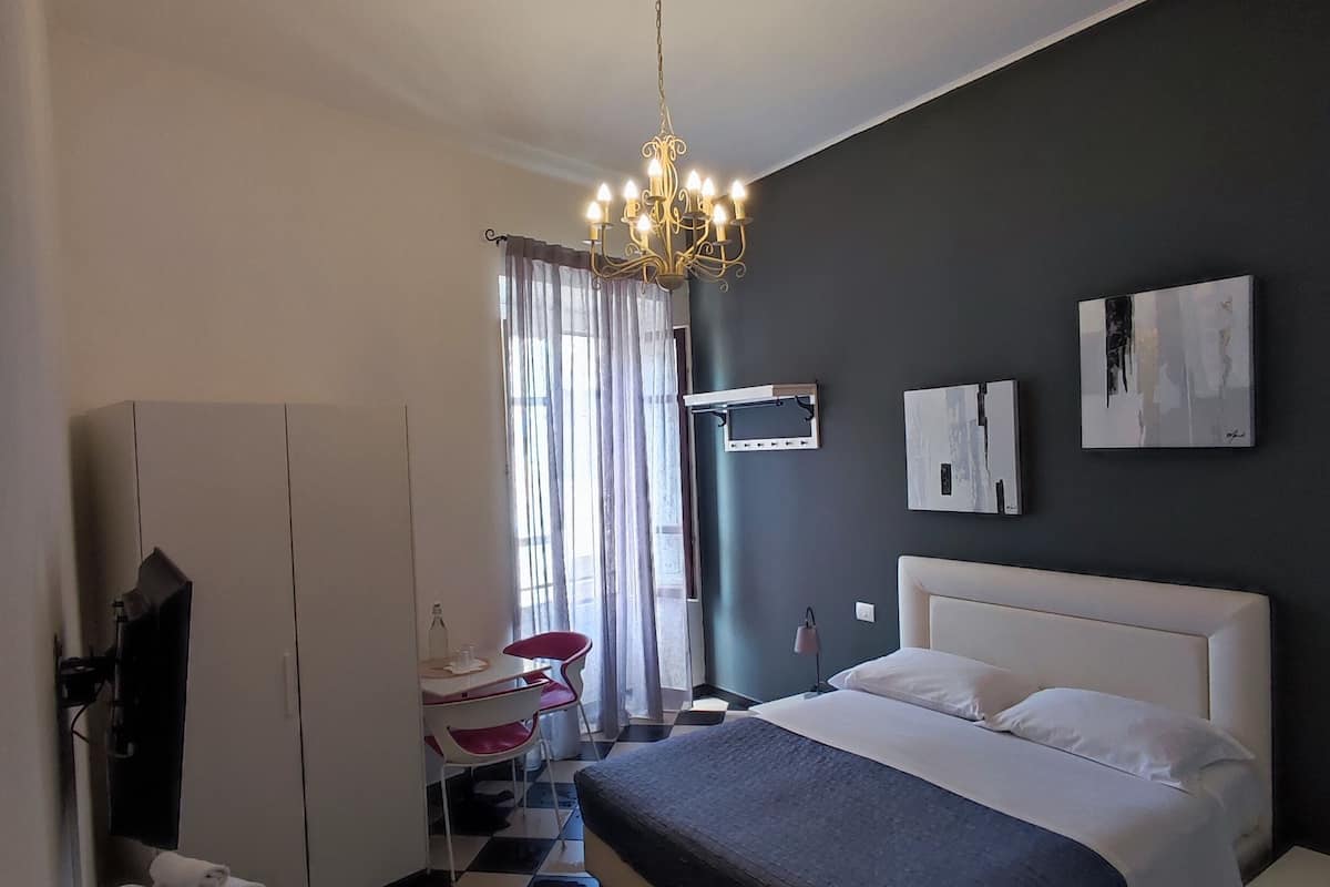 A hotel room with a checkered floor, a bed with white linens, a chandelier, a desk with a chair, a TV, and a window with curtains.