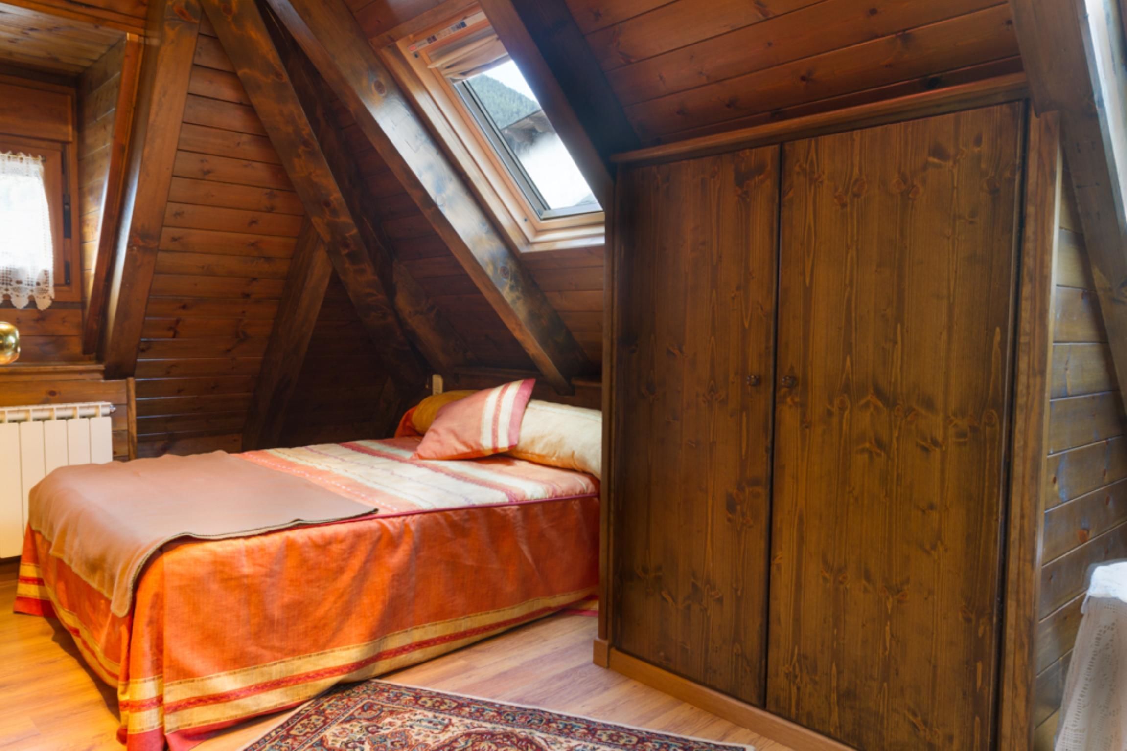Un dormitorio acogedor en el ático con techo inclinado, un ventanal grande, una cama con colcha colorida y un armario de madera.