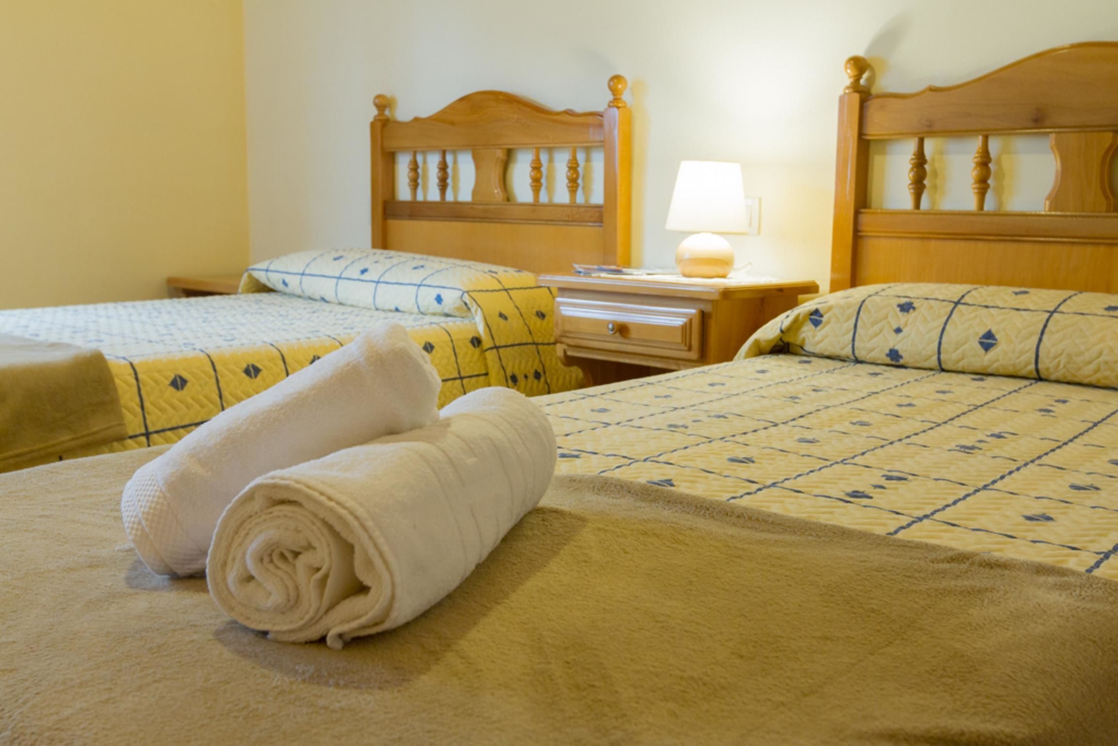 Habitación de hotel con dos camas, cada una con cabecero de madera, mesita de noche con lámpara y toallas ordenadamente dobladas.