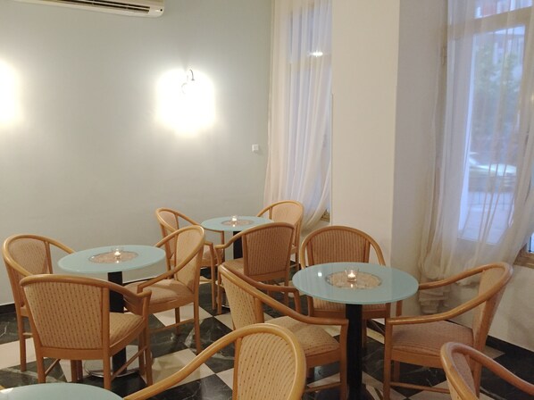 A room with wicker chairs and small round tables, a checkered floor, and a window with sheer curtains.
