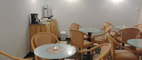 A dining area with round tables and wicker chairs, a coffee station, and wall-mounted lights.