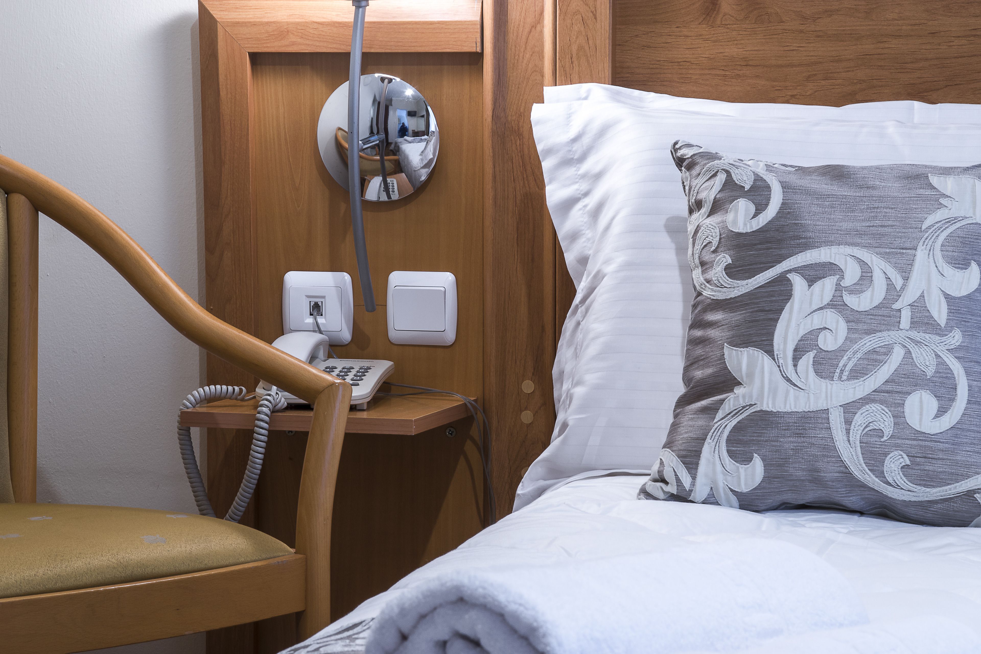 A hotel room with a wooden headboard, a bedside table with a telephone and a mirror, and a bed with white bedding and a decorative pillow.