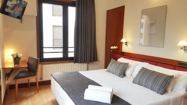 A hotel room with a bed, a chair, a desk, a window with curtains, and a wall-mounted TV.