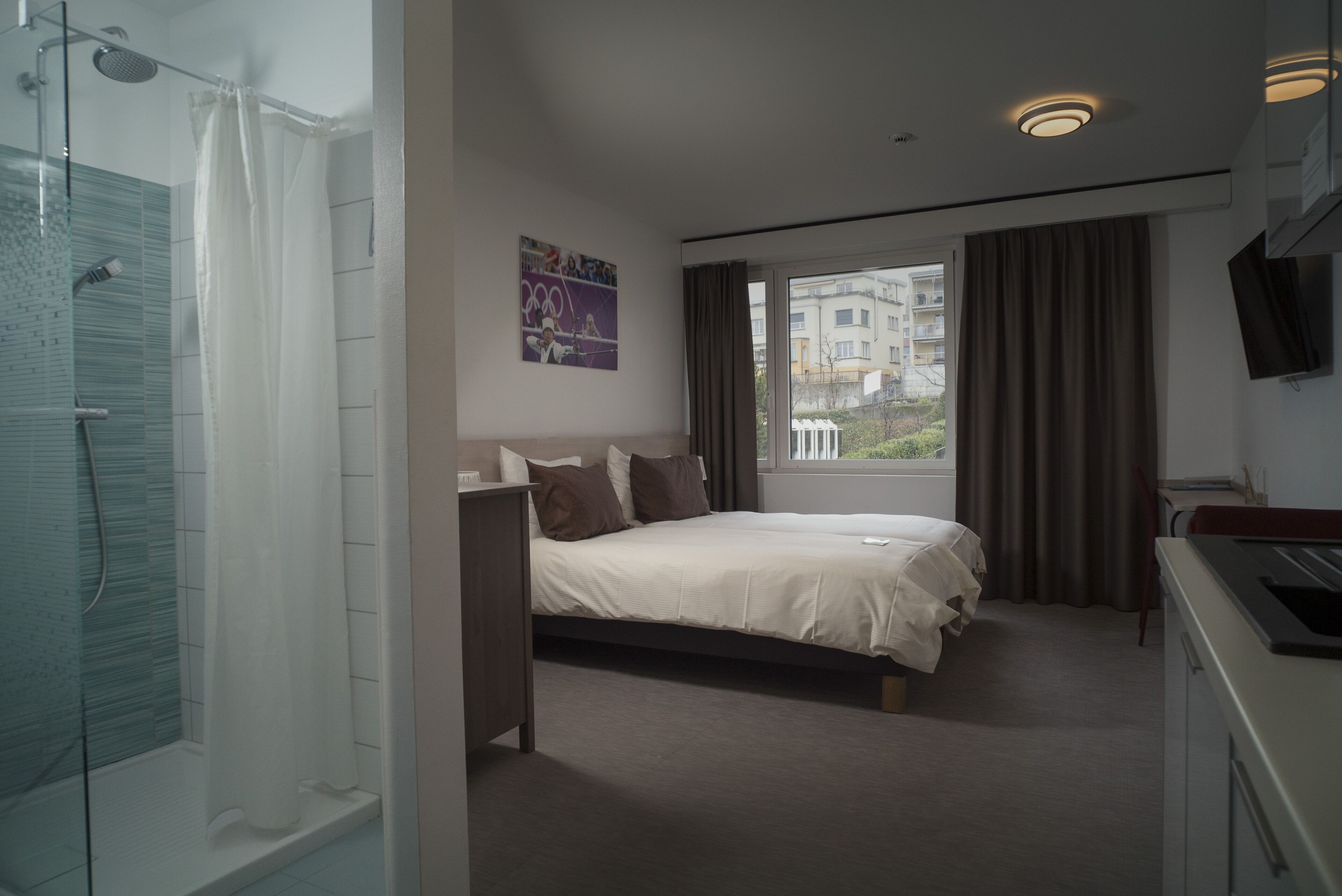 Ein Hotelzimmer mit Bett, Dusche, einem Fenster mit Vorhängen und einem Fernseher an der Wand.