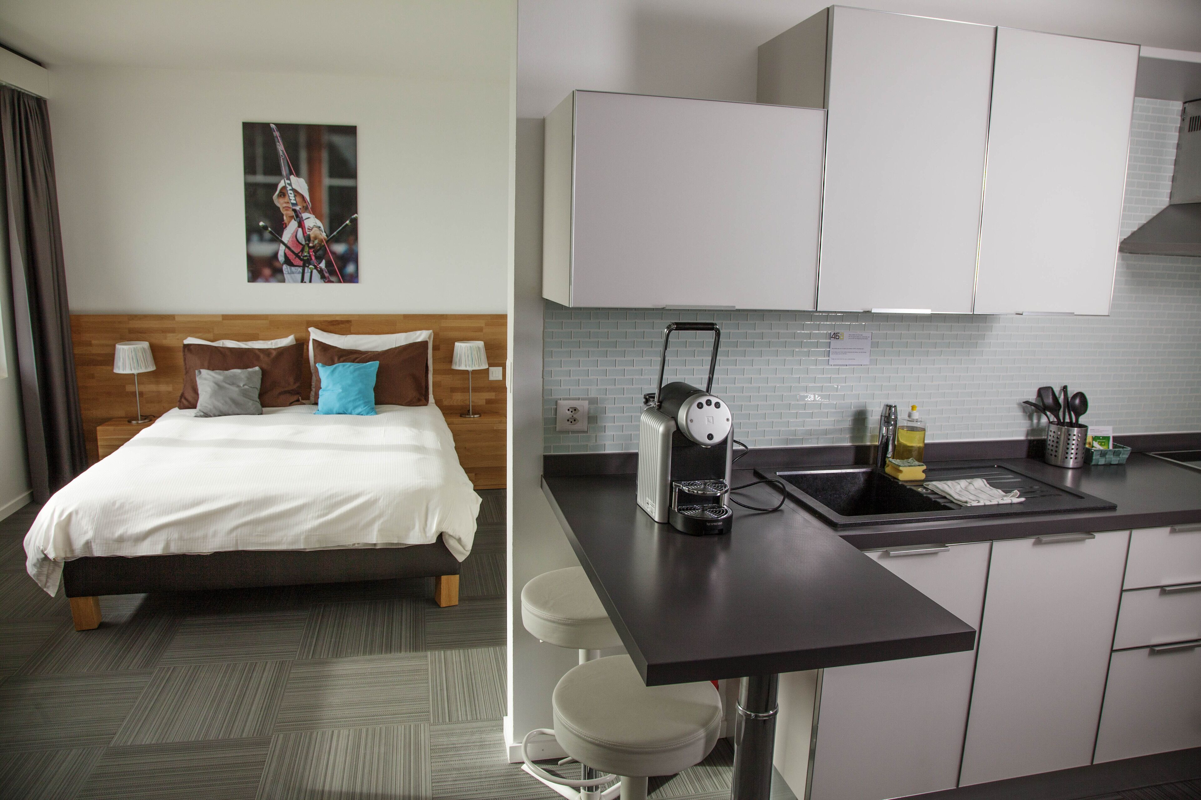 A hotel room with a bed, a nightstand, a lamp, and a picture on the wall. The room has a modern design with a wooden headboard and a grey carpet.