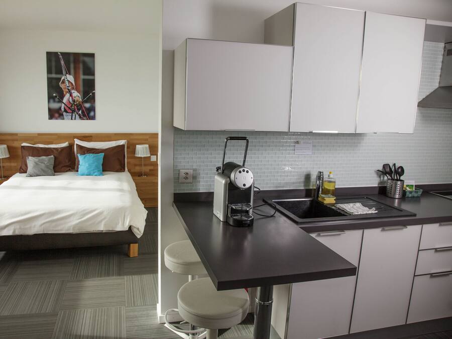 A hotel room with a bed, a nightstand, a lamp, and a picture on the wall. The room has a modern design with a wooden headboard and a grey carpet.