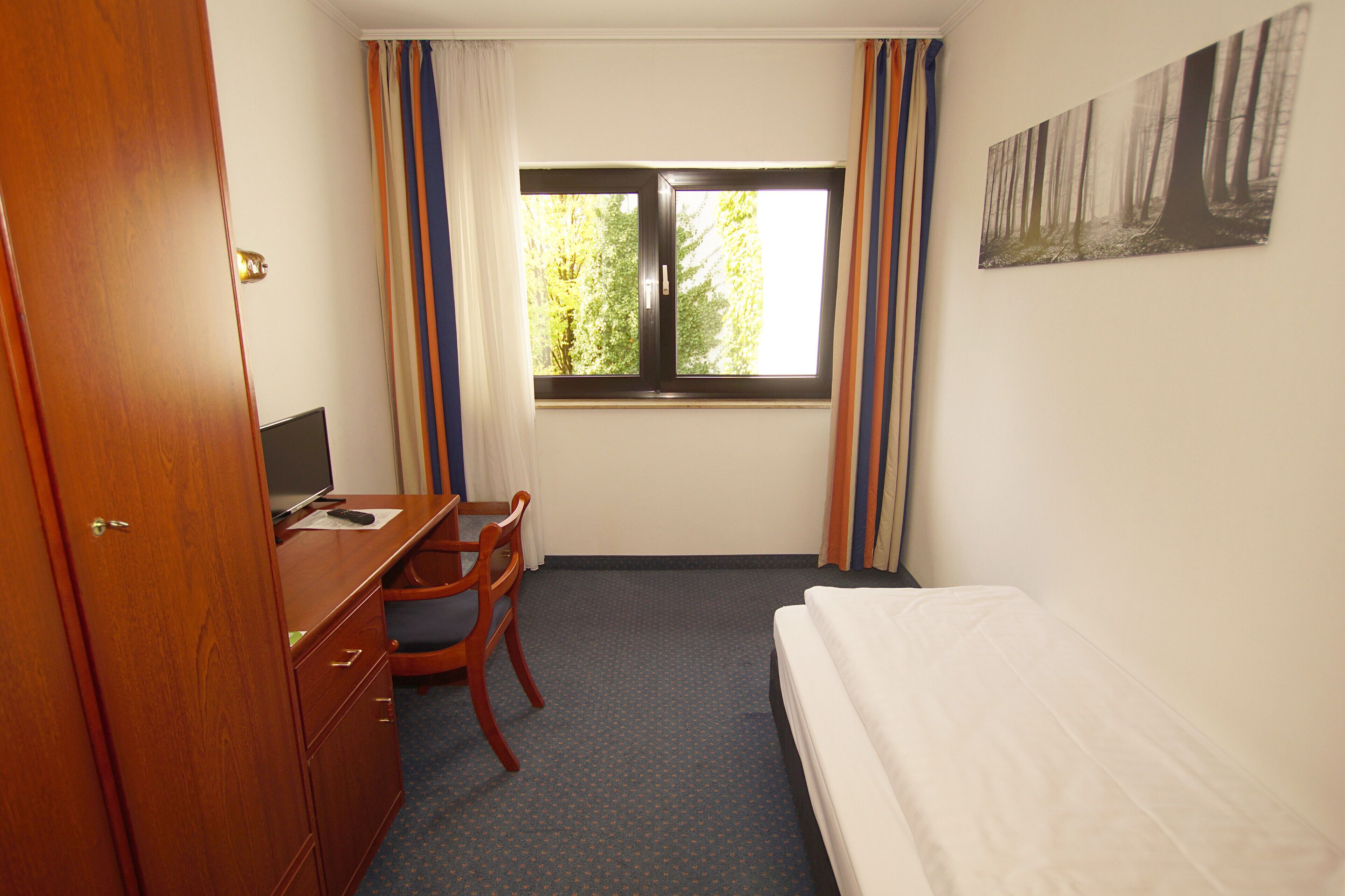 Habitación de hotel con cama, escritorio con silla, televisor y ventana con cortinas.