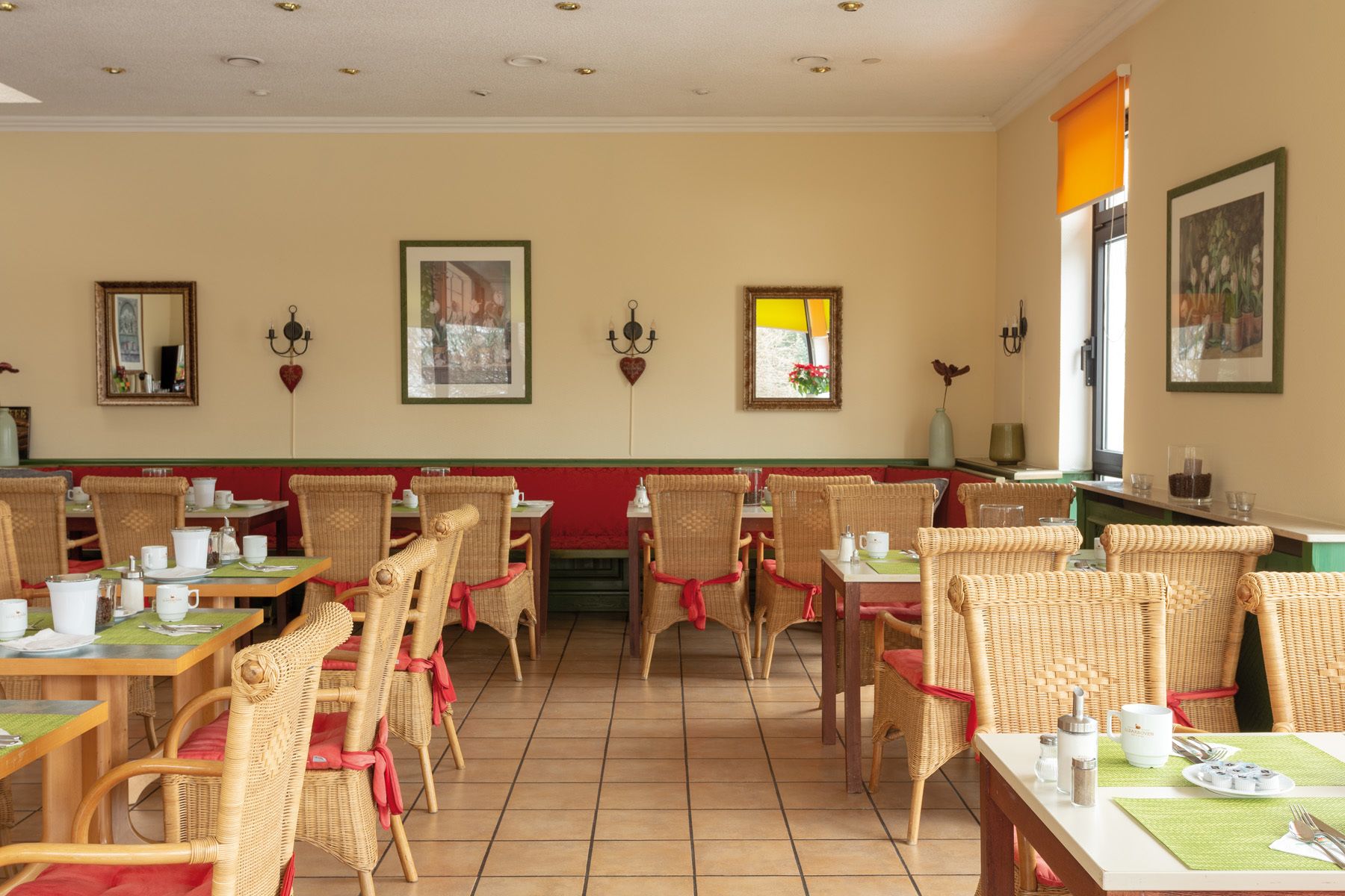 Een restaurant met rieten stoelen, groene tafelkleden en ingelijste foto’s aan de muur.