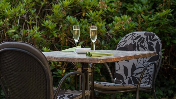 Een buitentafel met twee champagneglazen, een groene servet en een wit bord, voor een achtergrond van weelderige groene bladeren.