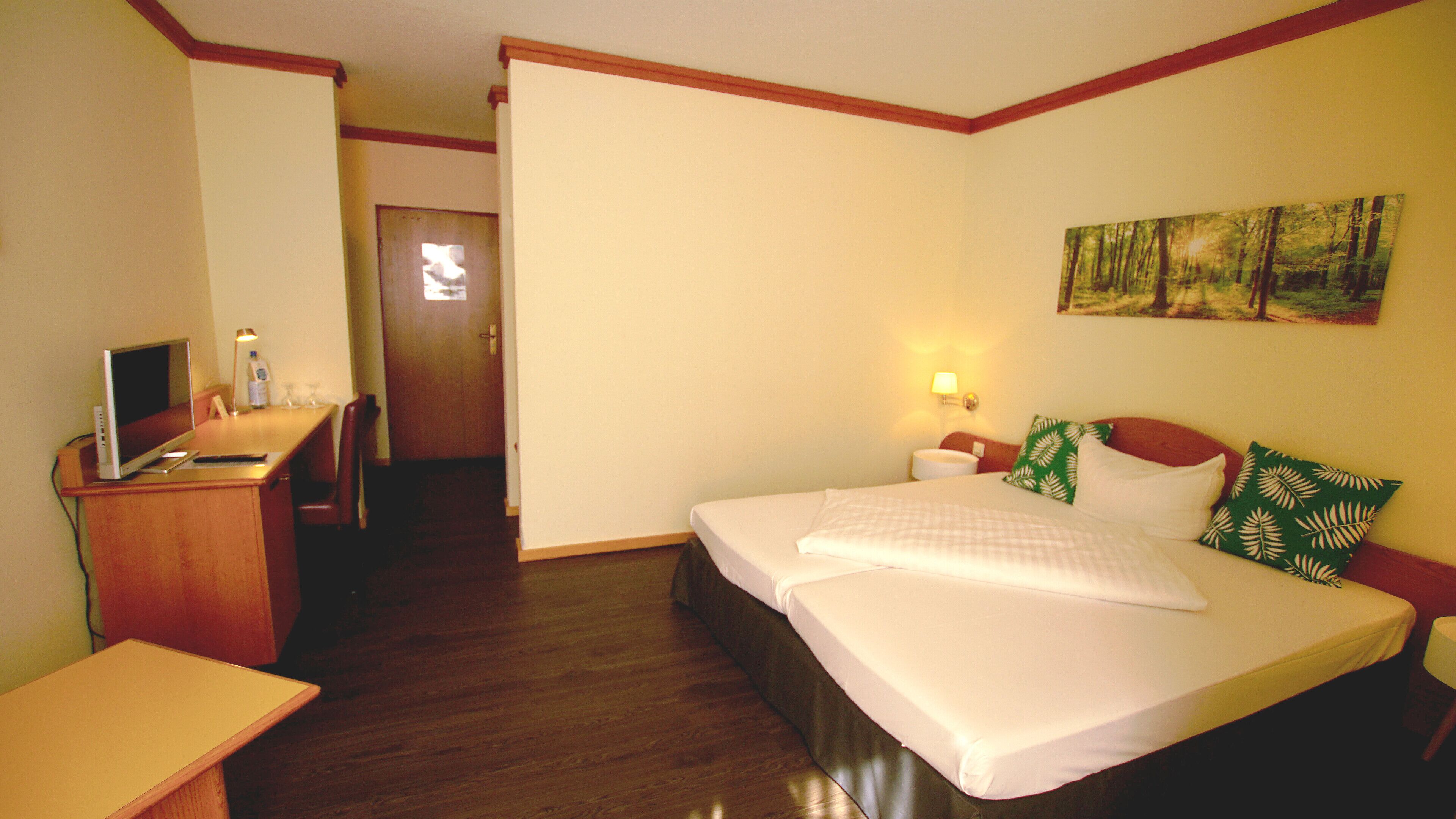 Habitación de hotel con una cama, un escritorio con computadora, una silla, una mesita de noche con lámpara y un cuadro en la pared.